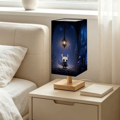 A decorative table lamp with a rectangular dark blue illustrated shade and a light wood base, placed on a bedside table next to a bed in a softly lit bedroom.