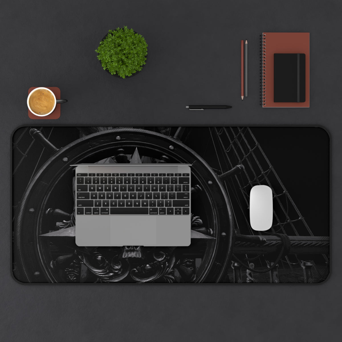 A large rectangular desk mat with a black and gray industrial wheel and machinery print, shown from above on a dark gray desk. A laptop, mouse, pen, notebook, and small plant are arranged around it as desk props. Assassin’s Creed black flag