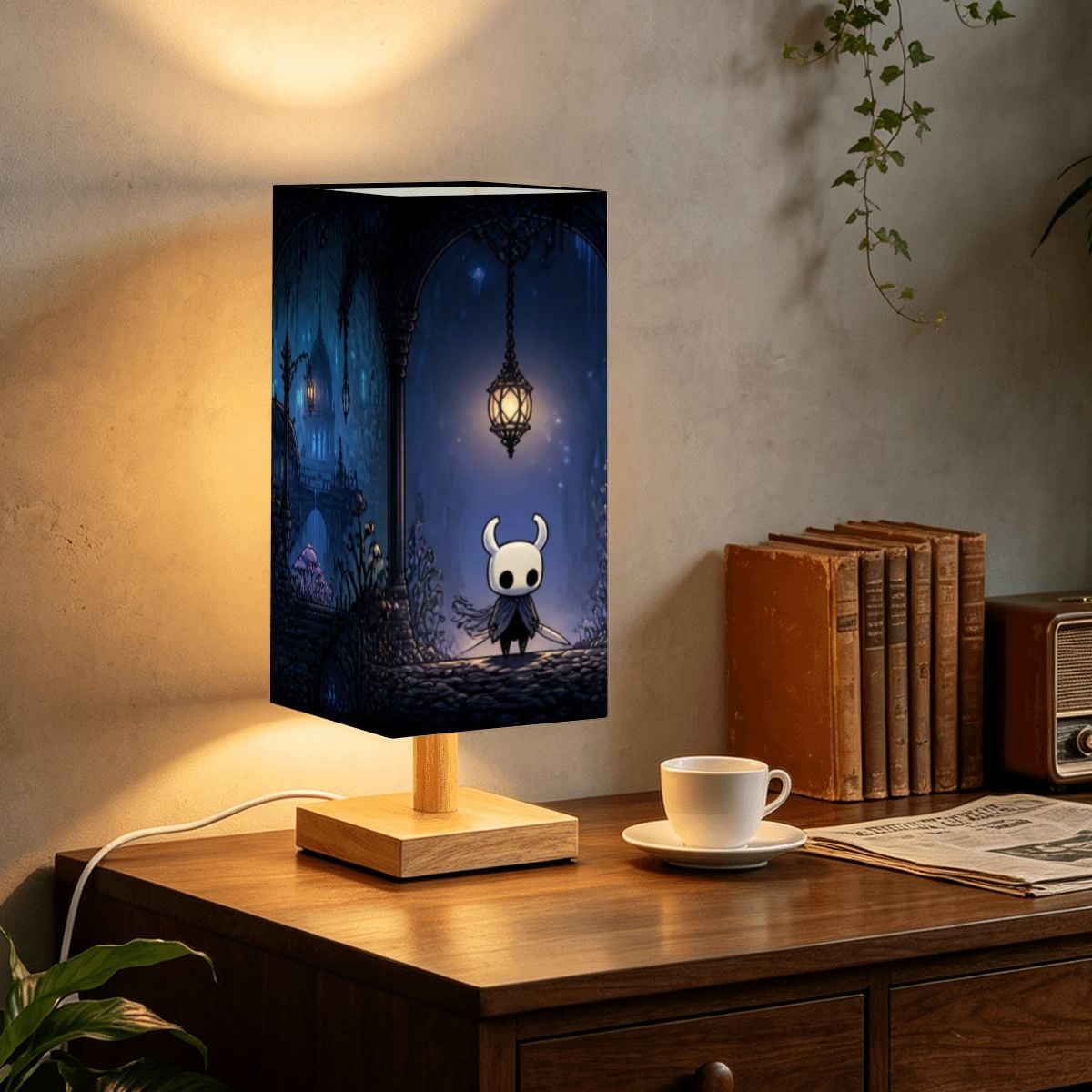 A rectangular table lamp with a printed fantasy scene on the shade sits on a wooden table in a cozy room setting. The lamp features a dark blue and black illustration with a small white character and a glowing lantern, supported by a light wood base.