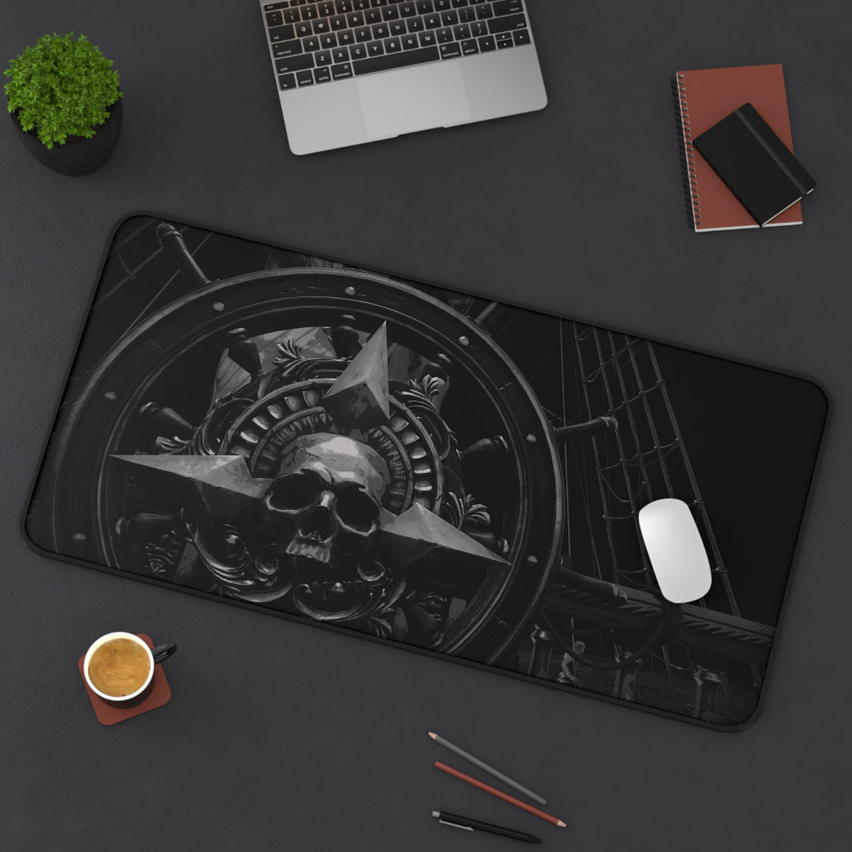 A large rectangular desk mat with a black and white gothic industrial skull graphic, styled on a dark gray desk with a laptop, mouse, coffee cup, and notebooks.