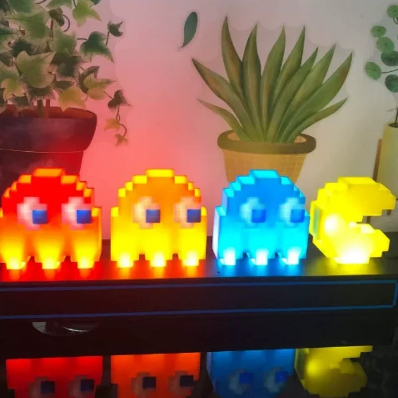 Four pixel-style ghost lights in red, yellow, blue, and green on a shelf with plants in the background.